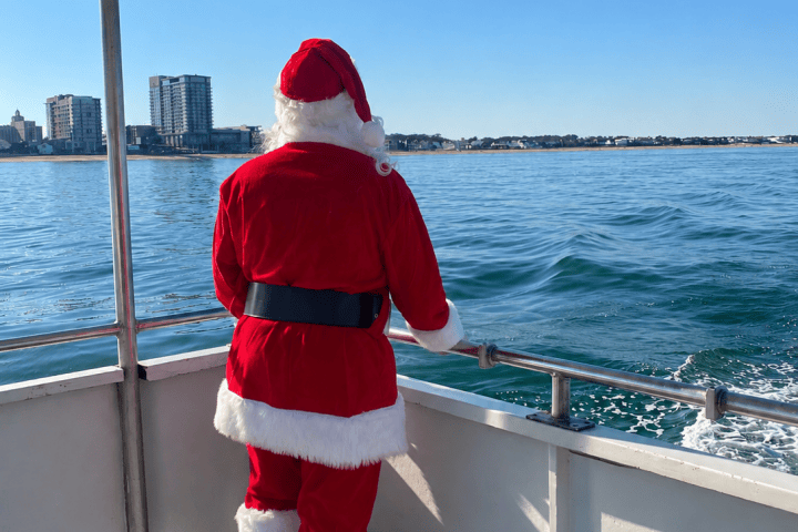 Santa standing on a boat, overlooking the water with city skyline in the distance.
