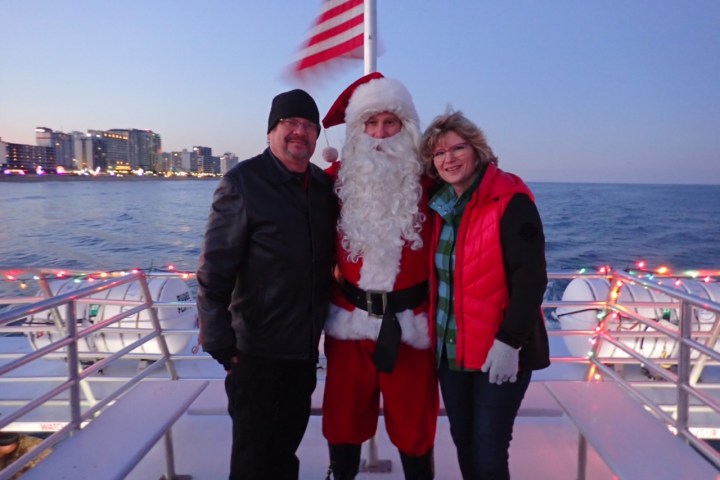 Three people, including one dressed as Santa, on a boat with a US flag and city skyline in the background.