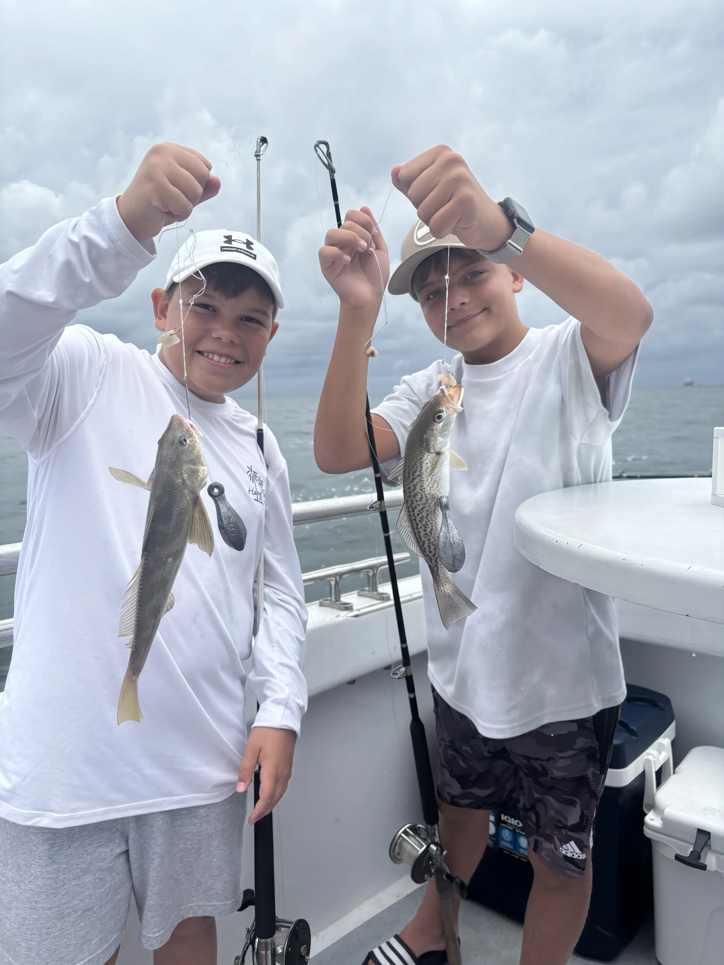 Croaker fishing. Half day trips. Two boys on a boat, holding fishing rods, each displaying a caught fish.
