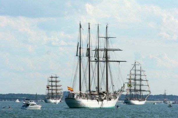 Tall ships and boats sailing on a calm sea under a cloudy sky.