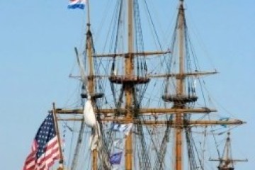 Historic tall ship sailing with multiple flags on a clear day.