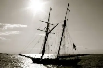 Silhouetted sailboat on water with sun overhead and flag visible.